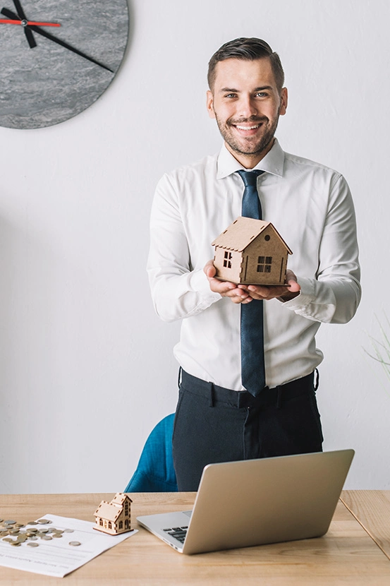 A person holding a model of a house standing behind a table with a laptop on it